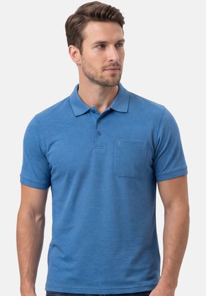 Mann trägt ein blaues Poloshirt mit kurzen Ärmeln und einer Brusttasche, schaut nach rechts vor einem schlichten Hintergrund.