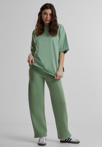 Žalia per didelė t-shirt ir atitinkančios plačios kojų kelnės iš minkštos medžiagos, su matomomis siūlėmis ir laisva iškirpte. Baltos sportbačiai.