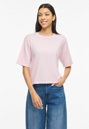 Kvinde smilende, iført en lyserød kortærmet skjorte og blå højtaljede jeans, stående med den ene hånd i lommen mod en hvid baggrund.