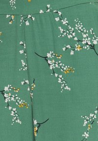 Grön tyg med ett blommönster i vitt och gula accenter, framhävt av svarta grenar. Slen struktur och lätt design.