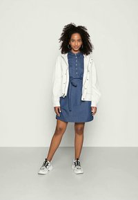 Vestido camiseiro em denim com botões à frente, cintura com cinto e camisolão branco. O modelo usa ténis preto e branco. O cabelo encaracolado realça o conjunto.