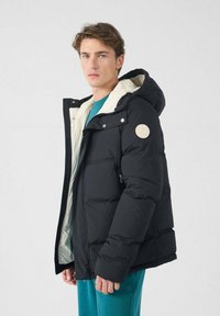 Zwarte pufferjack met een witte binnenvoering, capuchon, zijzakken en een ronde patch op de linkermouw. Gladde textuur, ritssluiting.