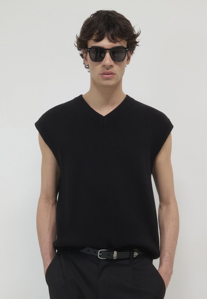 Schwarzer, gestrickter ärmelloser Pullover mit V-Ausschnitt und strukturiertem Finish, kombiniert mit schwarzen Sonnenbrillen. Das Model steht mit Händen in den Taschen vor einem weißen Hintergrund.