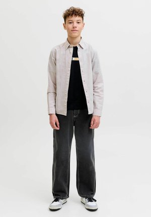 Teenager s kučeravými vlasmi má na sebe svetlobéžovú košeľu na gombíky cez čierne tričko s logom, voľné čierne rifle a biele tenisky, stojí.