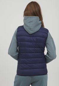 Navyblauwe gewatteerde vest met horizontaal gestikt ontwerp, gedragen over een lichtblauwe hoodiesweater. Gladde textuur, geïsoleerd voor warmte.
