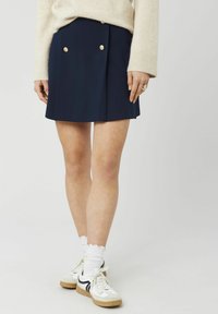 Jupe plissée marine avec des accents de boutons dorés, associée à des chaussettes blanches à volants et des baskets beiges avec des rayures noires. Texture de tissu lisse.