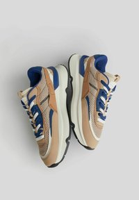 Pár béžových, modrých a bílých tenisek s síťovinou, silnými bílými podrážkami a modrými tkaničkami na světle šedém pozadí.