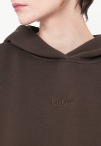 Hnedá mikina s jemnou textúrou, s veľkou kapucňou a vyšitým logom "LeGer" v mierne tmavšej hnedej farbe.