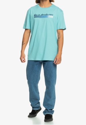 Camiseta de algodón turquesa con cuello redondo y mangas cortas; presenta un logo de Quiksilver en navy y un diseño de rayas horizontales en el pecho.