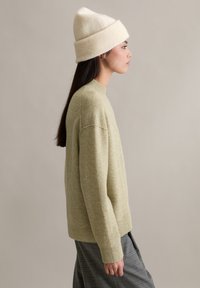 Pull tricoté vert clair à col rond, associé à un bonnet crème en maille côtelée. La silhouette porte une jupe grise à carreaux.