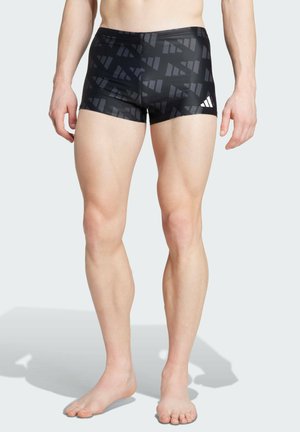 Shorts da bagno neri con un motivo geometrico, realizzati in materiale elastico. Presentano un design attillato e il logo Adidas in bianco sul lato.