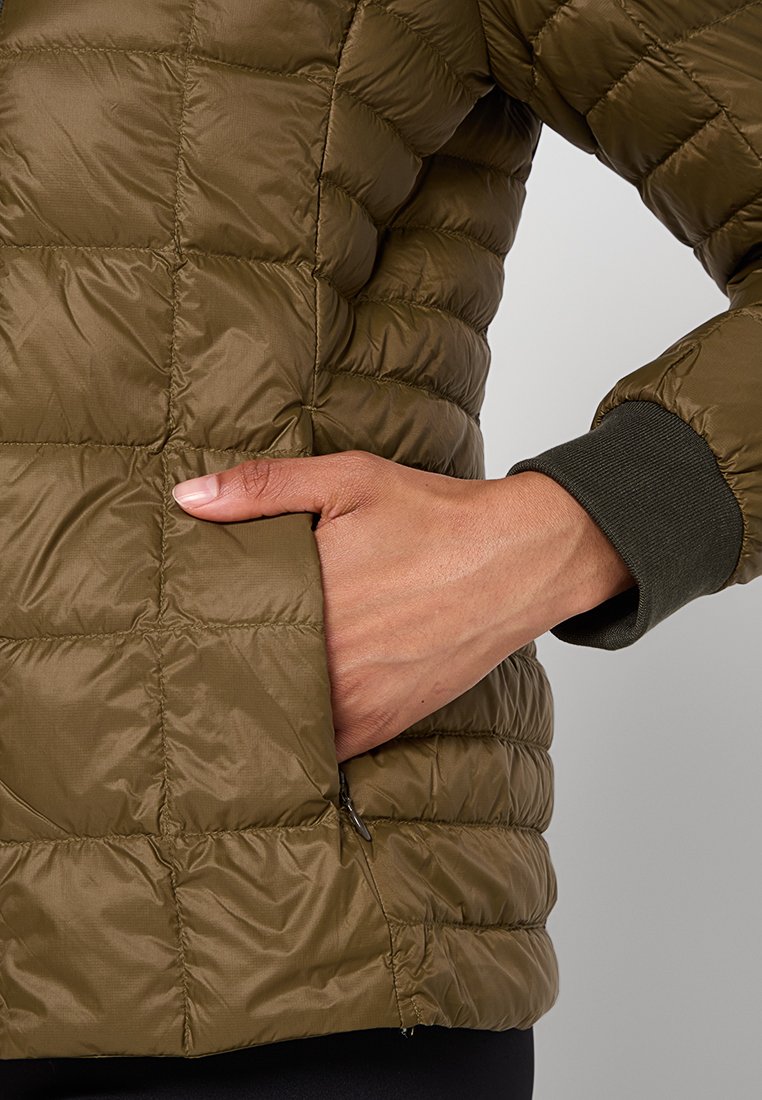 Rechte Hand in die mit einem Reißverschluss verschlossene Tasche einer olivgrünen gesteppte Jacke mit geripptem Bündchen gesteckt.