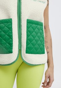 Chaleco de forro polar con bolsillos y ribetes acolchados verdes, cuerpo suave en crema, cierre con cremallera, costuras decorativas y diseño en capas.