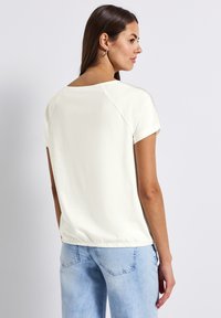 Top corto de manga corta en crema claro con escote redondeado, que presenta detalles texturizados en los hombros y un corte relajado, combinado con jeans azules.