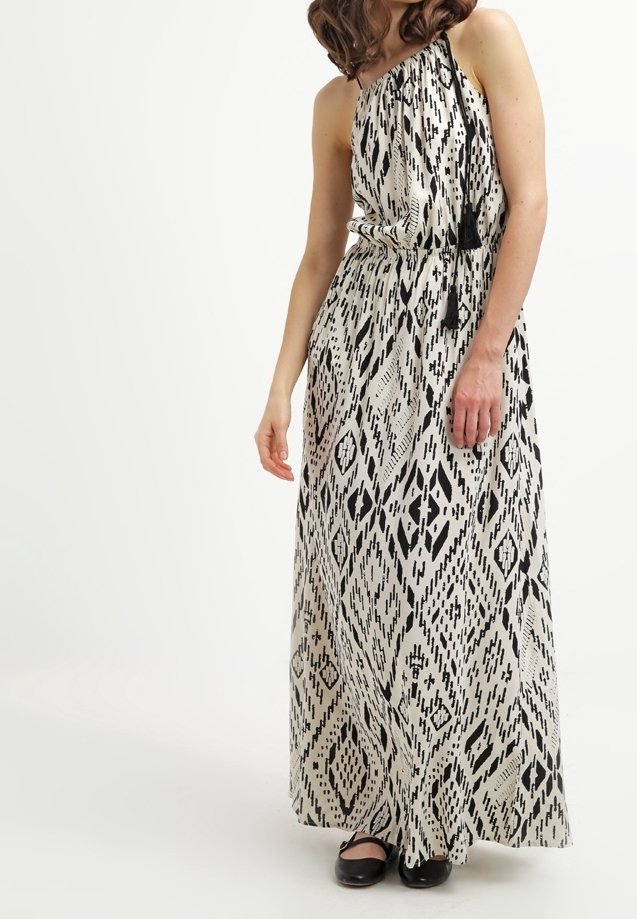 Femme portant une robe longue sans manches, à une épaule, noire et blanche à motifs, avec un lien à pompon noir et des chaussures plates noires.