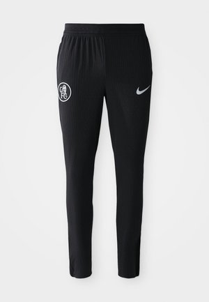 Pantalones deportivos negros con textura acanalada, que presentan un logo blanco de FC en la pierna izquierda y un swoosh gris de Nike en el lado derecho.