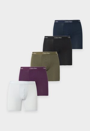 Mann steht barfuß da, trägt dunkelblaue Calvin Klein Boxershorts vor hellem, einfarbigem Hintergrund und schaut nach links.