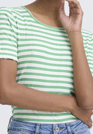 Personne portant une chemise à manches courtes rayée verte et blanche avec un jean en denim bleu, croisant un bras et touchant son visage avec l’autre main.
