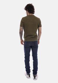 Homme aux cheveux bouclés et tatouages sur les bras portant un pull à manches courtes vert olive, un jean bleu et des baskets blanches, vu de dos.