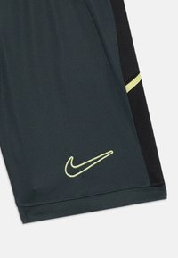 Nike šortai tamsiai pilko audinio su juodais tinklelio šonų elementais; turi geltoną logotipą ir kontrastingą geltoną juostą šone.