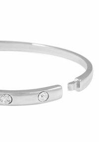 Sölj cuff-armband med två klara stenar, slät metallisk finish, öppen design med en liten lucka i den ena änden, minimalistisk stil.
