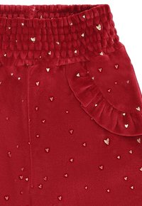 Pantaloni in velluto rosso con una cintura elastica testurizzata, caratterizzati da piccoli motivi a cuore dorato e un dettagli arricciato sulla tasca laterale.