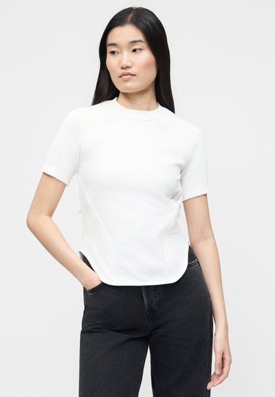 Νεαρή γυναίκα με μακριά μαύρα μαλλιά, ντυμένη με λευκό κοντομάνικο πλεκτό τοπ και μαύρο τζιν με ψηλή μέση, όρθια με το ένα χέρι στην τσέπη.