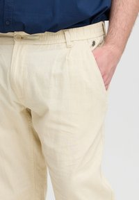 Lys beige bomullsbukser med elastisk linning, sidelommer og en glatt tekstur. Har en knappdetalj i midjen.