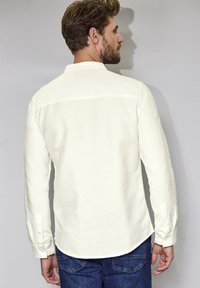 Langarmige, off-white shirt met een grof geweven stof, knoopmanchetten en een afgeronde zoom. Draag met blauwe jeans, weergegeven vanuit de rug.