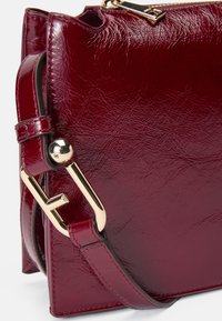 Borsa in pelle bordeaux con una texture liscia e lucida. Presenta dettagli in oro e un design strutturato con una tracolla removibile.