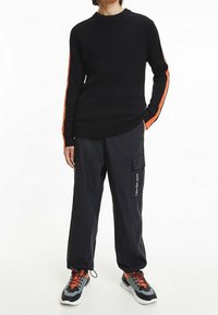 Schwarzer, gerippter Pullover mit orangenen Streifen, kombiniert mit schwarzen Cargohosen mit Seitentaschen und Calvin Klein Jeans Logo. Graue und orangefarbene Sneaker.