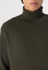 Pull à col roulé vert foncé avec un design texturé côtelé. Le tissu semble doux et épais, adapté aux temps plus frais.