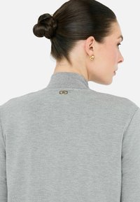 Felpa grigia chiara con colletto alto, texture liscia e un piccolo dettaglio in logo dorato sulla parte posteriore del collo. Design semplice e minimalista.