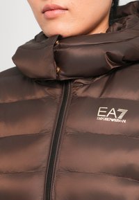 Hnedá puffová bunda s vysokým golierom, zipsovým zapínaním a logom EA7 Emporio Armani. Obsahuje prešívanú textúru a zlaté kovové doplnky.
