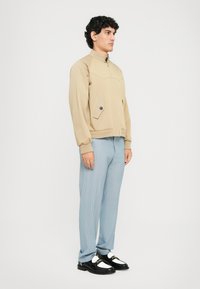 Bež bomber jakna z visokim ovratnikom, ki ima sprednji žep in gumbasti detajl. Svetlo modri hlače s črtami in črne čevlje.