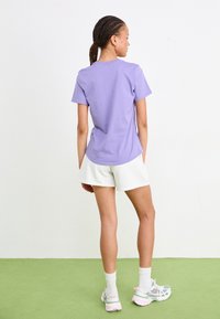 T-shirt violet à manches courtes avec un ourlet arrondi, associé à un short blanc. Le tissu semble lisse et confortable. L'arrière-plan est uni.
