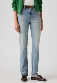 Φόρμα ανοικτού μπλε τζιν με ευθεία γραμμή, με ξεθωριασμένο φινίρισμα. Συνδυασμένο με πράσινη ζακέτα και καφέ παπούτσια με κορδόνια.