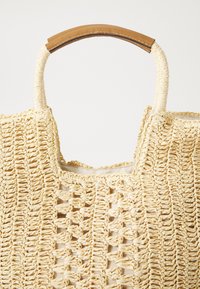 Sac en paille tissée avec un intérieur en tissu beige, doté d'une poignée courbe marron avec couture. Ouvert sur le dessus avec un motif texturé.