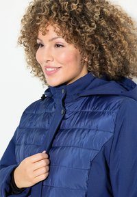 Giacca blu navy con una texture trapuntata sulla parte superiore, tessuto uniforme nella parte inferiore e cappuccio. Presenta una chiusura con bottoni e un cordino regolabile.