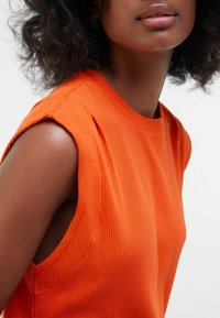 Top orange côtelé sans manches avec un col rond, présentant des coutures d'épaule subtiles et une texture lisse. Met en valeur la zone des bras.