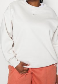 Ljusgrå sweatshirt med rund hals och lös passform, med en liten silverfärgad Nike-logga på bröstet och ribbade ärmslut, bärs tillsammans med korallfärgade shorts.