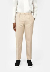 Beige skräddarsydda byxor med rak passform, utrustade med veck och sidofickor, paired med svarta sneakers. Outfitet inkluderar en randig skjorta.