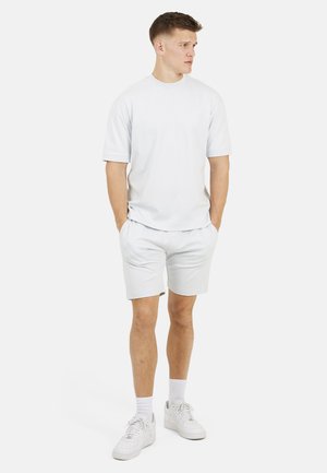 Młody mężczyzna stojący z rękami w kieszeniach, ubrany w całkowicie biały, codzienny strój, obejmujący T-shirt, szorty, białe skarpety i białe trampki.