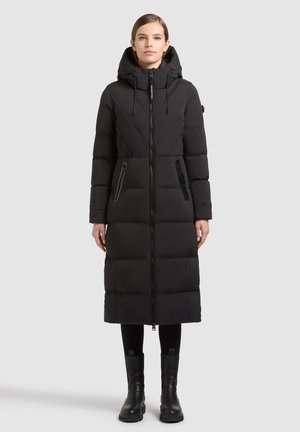 Femme portant un long manteau noir matelassé avec une capuche, fermé par une fermeture éclair, leggings noirs et bottines noires, debout sur un fond clair uni.