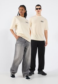 Beige overdimensjonerte t-skjorter på to modeller, en i grå jeans og den andre i svarte bukser, med sneakers. Enkelt tekst trykt på skjortene.