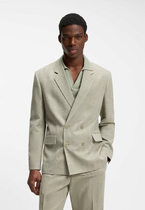 Jeune homme portant un blazer croisé beige clair et un pantalon assorti avec une chemise verte en dessous, debout devant un fond blanc.