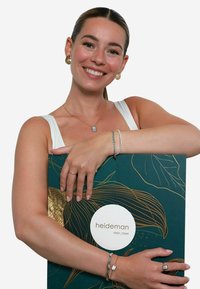 Cadeau vert et or en boîte avec un motif de feuilles et un logo rond, tenu par une personne portant des bijoux en argent et un haut blanc.