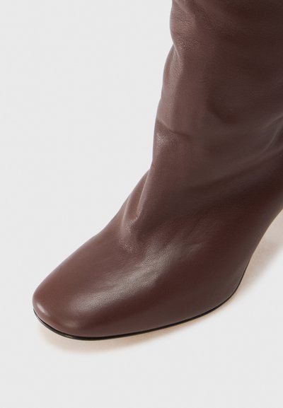 Brązowy skórzany botek za kostkę z cienkim, zaostrzonym czubkiem oraz teksturowaną, miękką cholewką. Na wysokim obcasie i z gładkim wykończeniem, na białym tle.