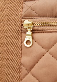 Primer plano de un tirador de cremallera YKK dorado sobre una tela acolchada beige, junto a un textil marrón con textura espiga.