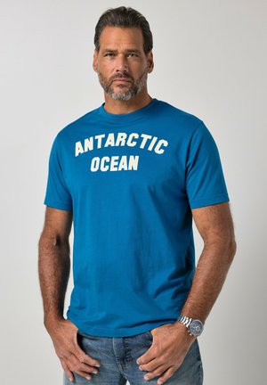 Mężczyzna z krótkimi ciemnymi włosami i brodą, ubrany w niebieską koszulkę z napisem „ANTARCTIC OCEAN” i zegarek na nadgarstku, stoi z rękami blisko kieszeni na szarym tle.
